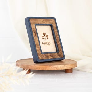 May include: A rectangular picture frame with a navy blue border and a wooden interior. The frame is leaning on a small, round wooden stand. The text "ARTSY FRAMES" is visible on the frame.