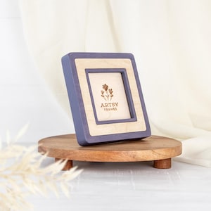 May include: A square picture frame with a light wood interior and a periwinkle blue exterior. The frame is resting on a small, round, wooden stand. The frame has the text "ARTSY FRAMES" and a floral design.