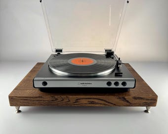 May include: An Audio-Technica turntable with a black chassis, silver front panel, and clear plastic dust cover, featuring a black vinyl record with an orange center label, resting on a rectangular dark wood platform with four small metal feet.