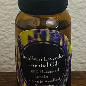 May include: A small brown glass bottle of ImaBean Lavender Essential Oils with a black cap. The label features a purple lavender design and text that reads "100% Phenomenal lavender oil Grown in Woodland Washington."