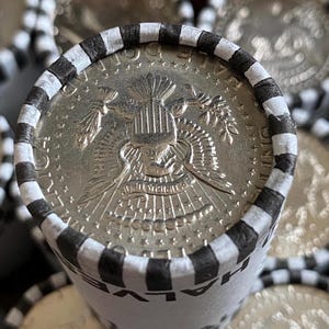 May include: A cylindrical paper roll containing United States half dollar coins. The top coin shows the presidential seal with an eagle, arrows, and olive branch. The paper wrapper is white with black stripes and the text HALF DOLLAR printed on it.