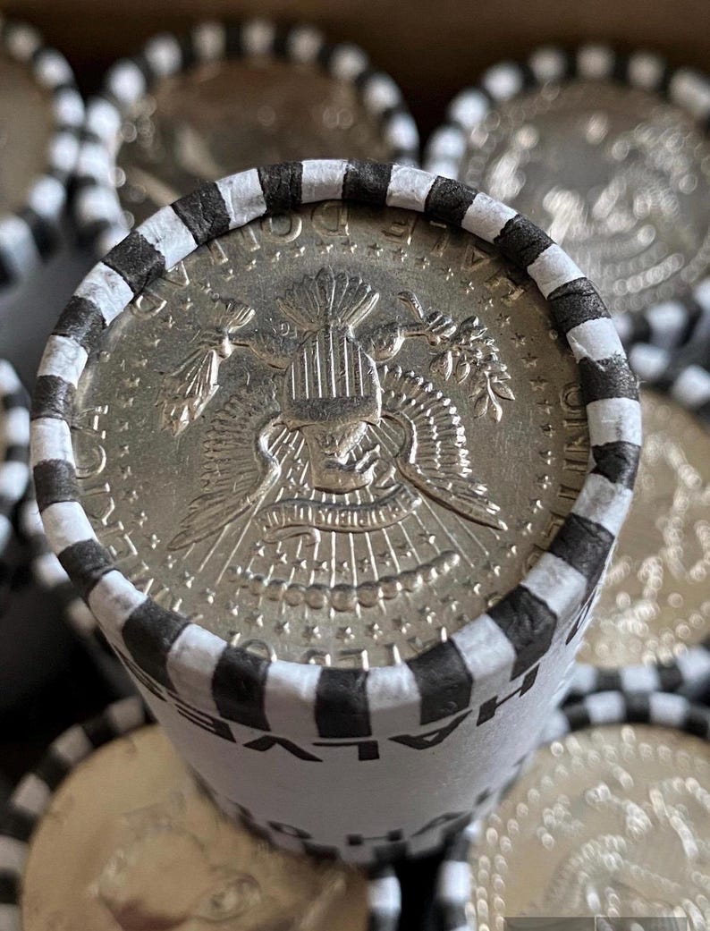 May include: A cylindrical paper roll containing United States half dollar coins. The top coin shows the presidential seal with an eagle, arrows, and olive branch. The paper wrapper is white with black stripes and the text HALF DOLLAR printed on it.