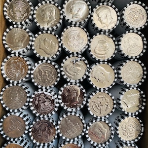 May include: A collection of rolled United States half-dollar coins arranged in a grid. Each paper roll is secured with black and white striped tape and displays either the profile of John F. Kennedy or the presidential seal on the visible end.