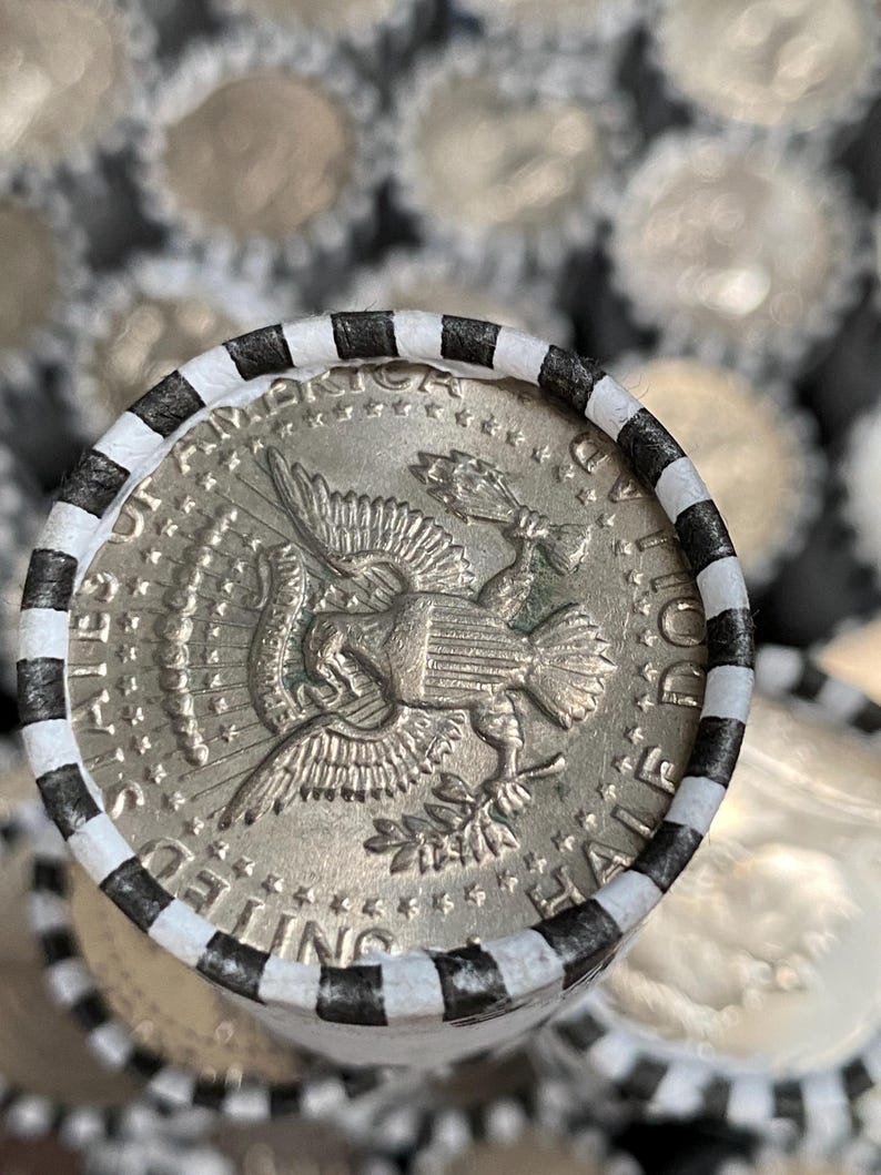 May include: A silver-colored United States half dollar coin featuring an eagle design on the reverse side, encased in a paper coin roll wrapper with alternating black and white stripes. The coin shows the text UNITED STATES OF AMERICA HALF DOLLAR.