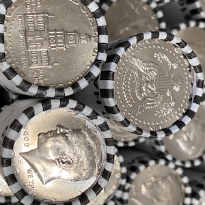 May include: Multiple rolls of silver-colored coins wrapped in white paper with black stripes. The visible coins show the profile of John F. Kennedy, the text IN GOD WE TRUST, and an engraving of Independence Hall, along with the presidential seal.