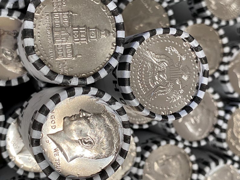 May include: Multiple rolls of silver-colored coins wrapped in white paper with black stripes. The visible coins show the profile of John F. Kennedy, the text IN GOD WE TRUST, and an engraving of Independence Hall, along with the presidential seal.