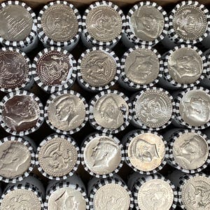 May include: A collection of silver-colored Kennedy half-dollar coins arranged in rows, each roll wrapped in white paper with black striped edges. The coins display the profile of John F. Kennedy or the presidential seal, showing metallic luster.