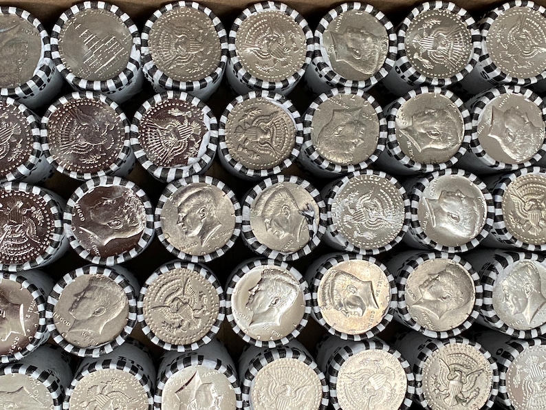 May include: A collection of silver-colored Kennedy half-dollar coins arranged in rows, each roll wrapped in white paper with black striped edges. The coins display the profile of John F. Kennedy or the presidential seal, showing metallic luster.
