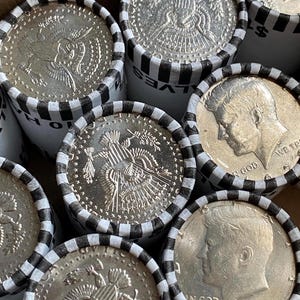 May include: Multiple rolls of United States half-dollar coins wrapped in white paper with black striped edges. The visible coins display the profile of John F. Kennedy and the Presidential Seal, with text including IN GOD WE TRUST and HALVES.