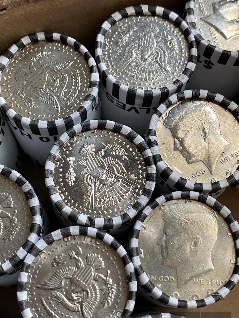 May include: Multiple rolls of United States half-dollar coins wrapped in white paper with black striped edges. The visible coins display the profile of John F. Kennedy and the Presidential Seal, with text including IN GOD WE TRUST and HALVES.