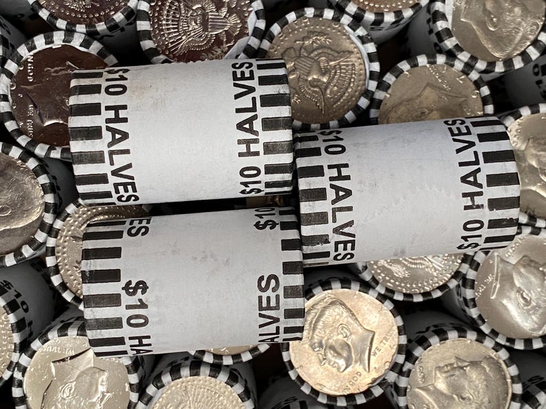 May include: Multiple cylindrical paper coin rolls filled with United States half-dollar coins. Each white wrapper features black vertical stripes and the printed text $10 HALVES. The ends of the rolls reveal the metallic silver surfaces of the coins.
