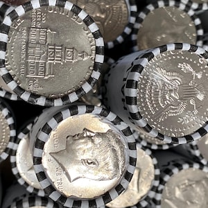 May include: A collection of rolled United States half-dollar coins. Each roll is secured with white paper wrappers featuring black stripes. The visible coins display the profile of John F. Kennedy, the Presidential Seal, and Independence Hall.