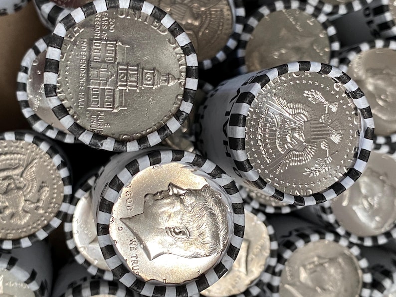 May include: A collection of rolled United States half-dollar coins. Each roll is secured with white paper wrappers featuring black stripes. The visible coins display the profile of John F. Kennedy, the Presidential Seal, and Independence Hall.