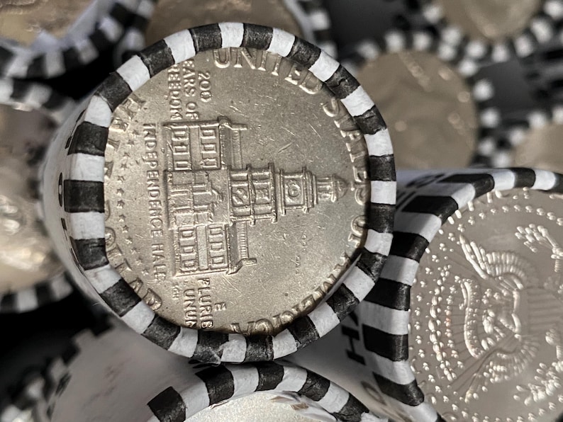 May include: A silver-colored United States half dollar coin featuring Independence Hall, encased in a white paper coin roll wrapper with black stripes. The coin shows the text 200 YEARS OF FREEDOM, INDEPENDENCE HALL, and E PLURIBUS UNUM.