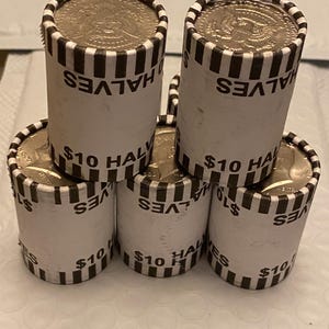 May include: Five cylindrical paper rolls of United States half-dollar coins stacked in a pyramid formation. Each white wrapper features black vertical stripes at the top and bottom edges, with the printed text $10 HALVES visible on each roll.