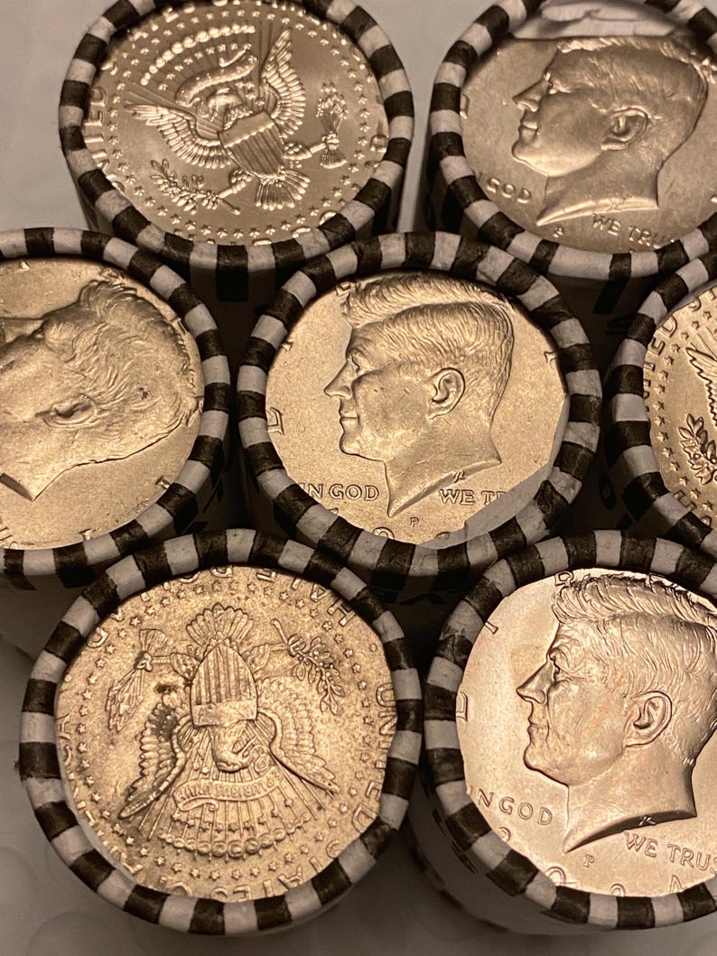 May include: Several rolls of United States half-dollar coins wrapped in white paper with black stripes. The visible coins feature the profile of John F. Kennedy and the presidential seal, with the text IN GOD WE TRUST clearly visible on the faces.