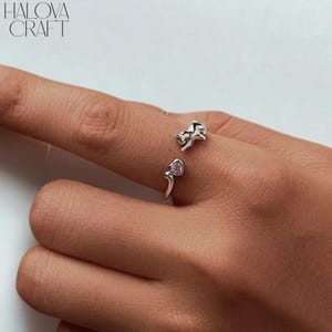 Könnte beinhalten: Ein silberner Ring mit einer Katze und einem herzförmigen rosa Edelstein. Der Ring hat ein offenes Design, wobei sich Katze und Herz auf gegenüberliegenden Seiten befinden. Der Markenname "HALOVA CRAFT" ist oben links sichtbar.