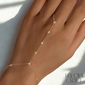 Puede incluir: Una delicada cadena de mano dorada con brillantes piedras preciosas transparentes. La cadena conecta un anillo con una pulsera, con pequeñas gemas redondas y en forma de flor. La joya se muestra en una mano, con la marca "HALOVA CRAFT" visible.