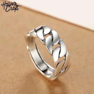 Könnte beinhalten: Ein silberfarbener Ring mit einem Kettenglied-Design. Der Ring ist verstellbar und verfügt über eine Reihe von miteinander verbundenen Gliedern, die ein kühnes und stilvolles Accessoire schaffen. Der Ring wird auf einer braunen Oberfläche präsentiert. Der Markenname "Halova Craft" befindet sich oben links.