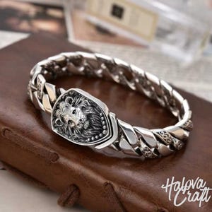 Könnte beinhalten: Silberfarbenes Armband mit einem Löwenkopf-Emblem. Das Armband hat ein dickes Kettendesign mit einem detaillierten Löwenkopf-Mittelstück. Das Armband wird auf einer braunen Lederoberfläche präsentiert. Der Text "Halova Craft" ist sichtbar.