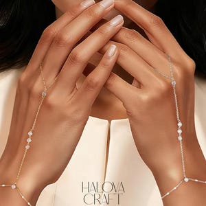 Op de afbeelding: Gouden en zilveren handketting sieraden met heldere edelstenen. De sieraden verbinden de vinger met de pols. De merknaam "HALOVA CRAFT" wordt weergegeven in witte tekst.