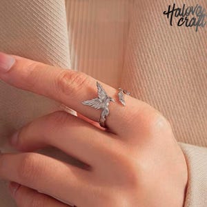 Könnte beinhalten: Ein silberner Ring mit einem detaillierten Vogeldesign und ausgebreiteten Flügeln. Ein Federdetail befindet sich ebenfalls am Ring. Der Ring umschließt den Finger und ist ein einzigartiges Schmuckstück.
