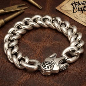May include: A polished silver curb chain bracelet featuring thick, rounded interlocking links. The bracelet includes a secure, heavy-duty lobster claw clasp with a circular floral cutout design and engraved text on the metal surface.