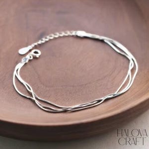 May include: A silver bracelet with three fine strands, displayed on a wooden surface. The bracelet has a small clasp and an adjustable chain. The brand name "HALOVA CRAFT" is visible in the bottom right corner.
