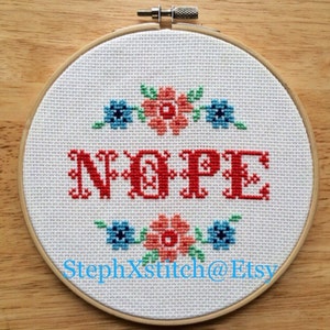 May include: A white embroidery hoop with a red cross-stitch saying "NOPE" surrounded by blue and orange flowers. The hoop is on a wooden surface. The text "StephXStitch@Etsy" is in the bottom right corner.