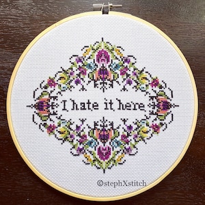 May include: A white embroidery hoop with a cross-stitch design featuring a floral pattern and the text "I hate it here".