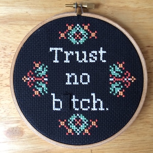 May include: A black cross-stitch embroidery hoop with the text "Trust no b*tch." in white. The design includes four decorative floral patterns in green, orange, and white. The hoop is set in a wooden frame.
