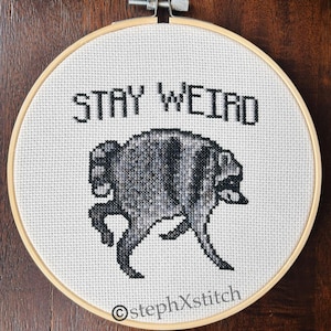 Könnte beinhalten: Ein runder Stickrahmen aus Holz mit weißem Aida-Stoff, der ein Kreuzstichmotiv einer runden, grau-schwarzen, waschbärähnlichen Kreatur zeigt. Der Text STAY WEIRD ist in Schwarz darüber gestickt, und stephXstitch befindet sich am unteren Rand.