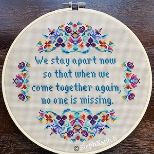 May include: A cross-stitch embroidery hoop with the text "We stay apart now so that when we come together again, no one is missing." The design features floral patterns in teal, purple, red, and yellow. The hoop is set against a neutral fabric.