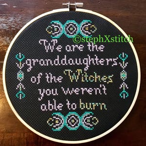 May include: A black embroidery hoop with a white frame, featuring a cross-stitch design with the text "We are the granddaughters of the Witches you weren't able to burn." The design also includes three triple moon symbols.