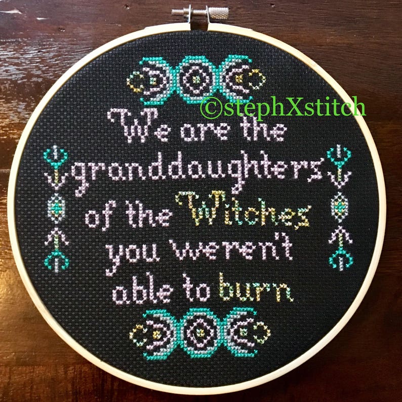May include: A black embroidery hoop with a white frame, featuring a cross-stitch design with the text "We are the granddaughters of the Witches you weren't able to burn." The design also includes three triple moon symbols.