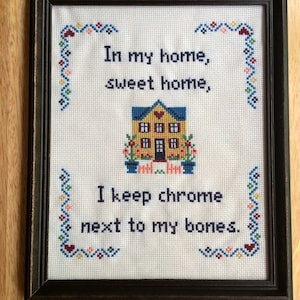 May include: A framed cross-stitch with a yellow house and the text "In my home, sweet home, I keep chrome next to my bones."