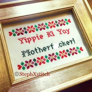 Könnte beinhalten: Kreuzstich-Kunstwerk in einem goldenen Rahmen. Der Text lautet "Yippie Ki Yay Motherf*cker!" in Rot und Schwarz. Florale Bordüren in Rot, Grün und Gold akzentuieren den Text. Der Name des Künstlers, StephXstitch, befindet sich unten links.