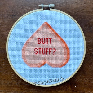 May include: A light blue cross-stitch embroidery hoop with a peach-colored heart shape in the center. The heart has the text "BUTT STUFF?" in red lettering. The hoop has the text "@StephXStitch" in the bottom right corner.