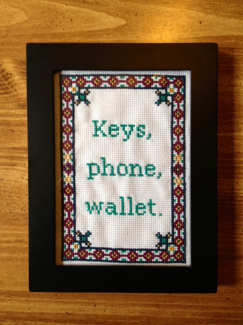 May include: A framed cross-stitch with a white background and a red, gold, and black border. The cross-stitch says "Keys, phone, wallet." in green thread.