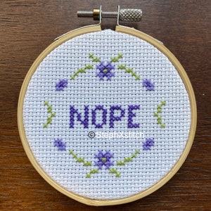 May include: A cross-stitch embroidery hoop displaying the word "NOPE" in purple thread. The design features purple flowers and green vines. The wooden hoop is secured with a metal screw. The text "StephXstitch" is also visible.