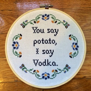 Op de afbeelding: Witte borduurring met een bloemrand en de tekst "You say potato, I say vodka."