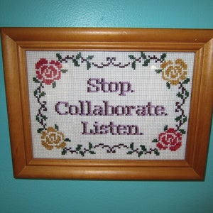 May include: A framed cross-stitch with a white background and a purple message that reads "Stop Collaborate and Listen." The message is surrounded by a border of green vines and colorful flowers.