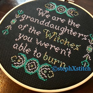 May include: A black embroidery hoop with a white and pink cross-stitch design. The design features a triple moon symbol and the text "We are the granddaughters of the Witches you weren't able to burn". The text is in a cursive font and the triple moon symbol is in a stylized design. The hoop is surrounded by a wooden frame.