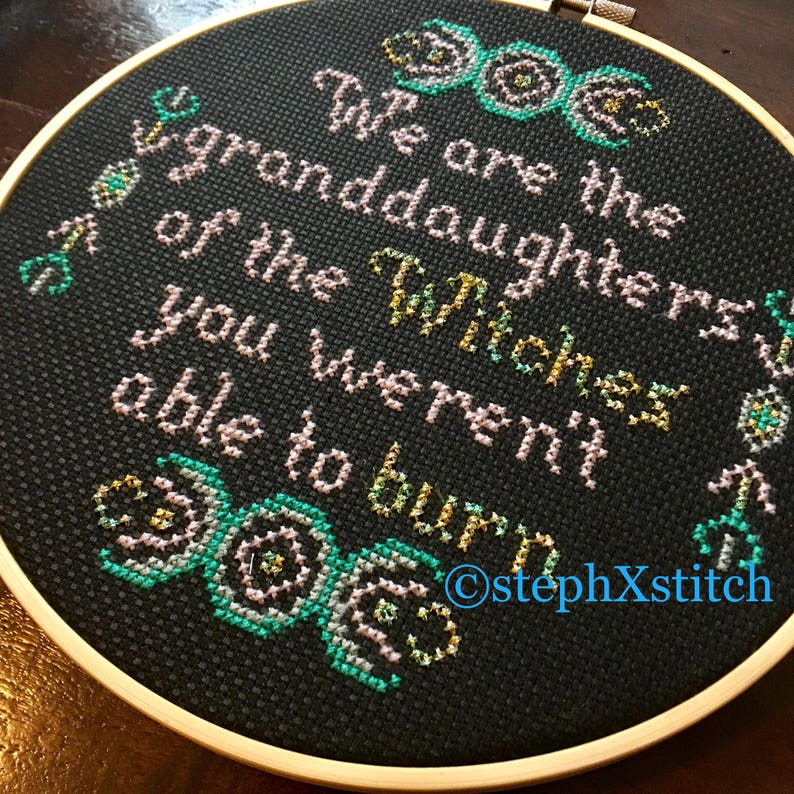 May include: A black embroidery hoop with a white and pink cross-stitch design. The design features a triple moon symbol and the text "We are the granddaughters of the Witches you weren't able to burn". The text is in a cursive font and the triple moon symbol is in a stylized design. The hoop is surrounded by a wooden frame.