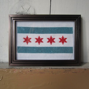 May include: A framed cross-stitch of the Chicago flag. The flag is white with six horizontal blue and white stripes. There are four red stars in the center of the flag.