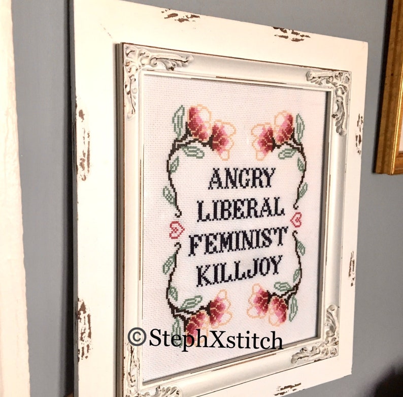 May include: White framed cross-stitch embroidery with pink flowers and the text "Angry Liberal Feminist Killjoy" and the copyright symbol and "StephXStitch".