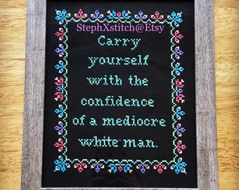 May include: Cross-stitch art in a gray wooden frame. The black fabric features the text "Carry yourself with the confidence of a mediocre white man" in turquoise letters. The border has a floral design in pink, blue, and green.