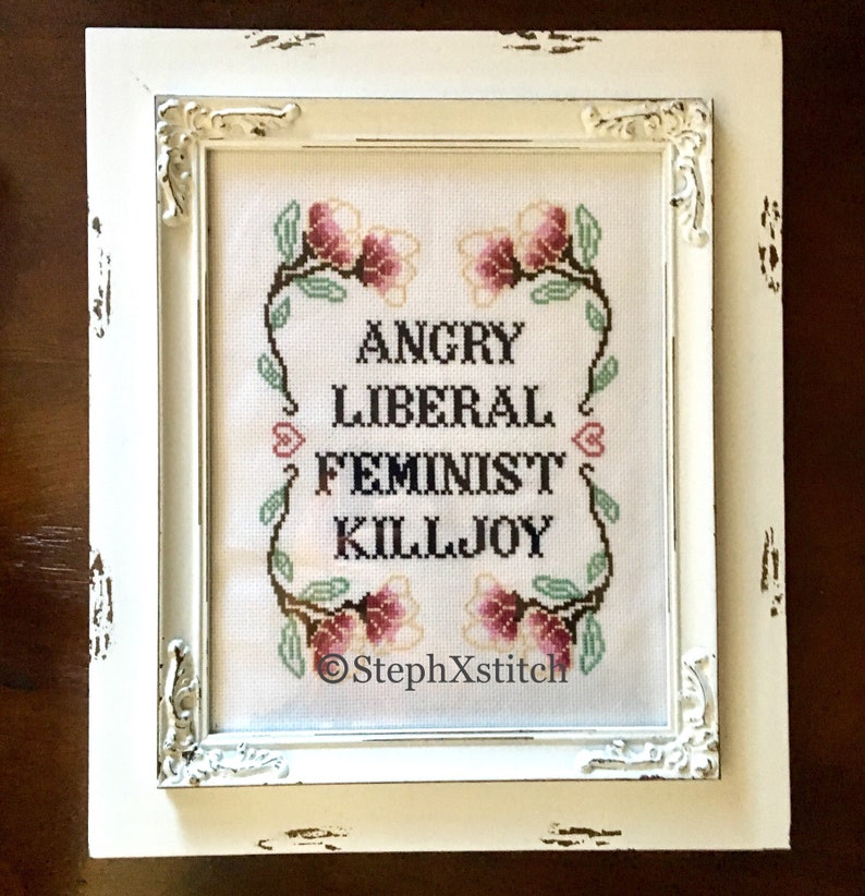 May include: White framed cross-stitch embroidery with pink flowers and the text "Angry Liberal Feminist Killjoy" and the signature "StephXStitch".