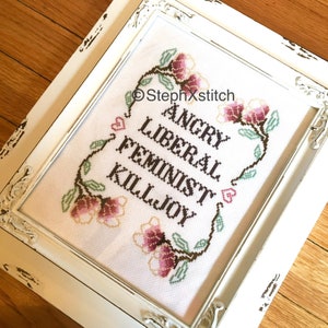 May include: A white framed cross-stitch with a floral design and the text "Angry Liberal Feminist Killjoy" in brown lettering.