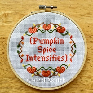 May include: A white embroidery hoop with a cross-stitch design featuring a vine of orange pumpkins and the text "(Pumpkin Spice Intensifies)". The hoop is made of wood and has a natural finish. The text "stephXstitch" is also included in the design.
